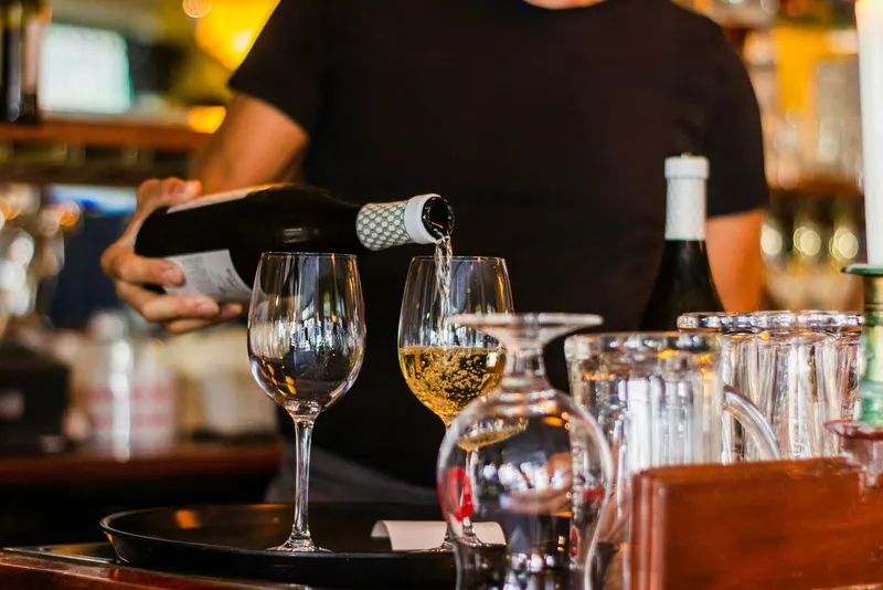 Un serveur en T-shirt noir verse du vin blanc dans un verre au bar d’un restaurant